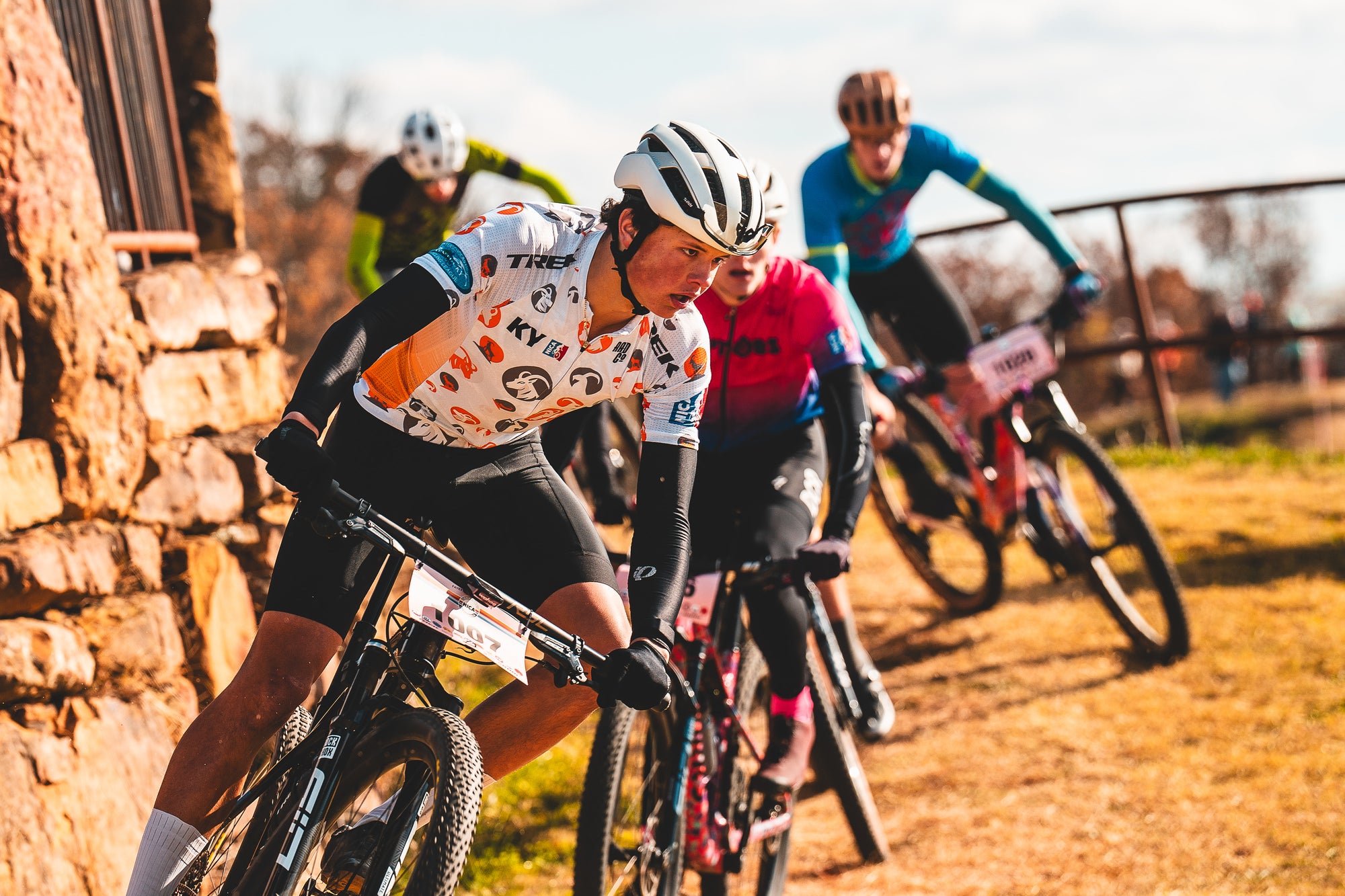 Cyclists racing on a dirt track with a stone wall in the background